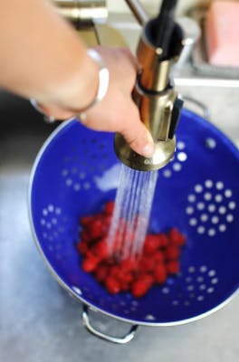 A hand holding a faucet spraying water over berries in a blue colander