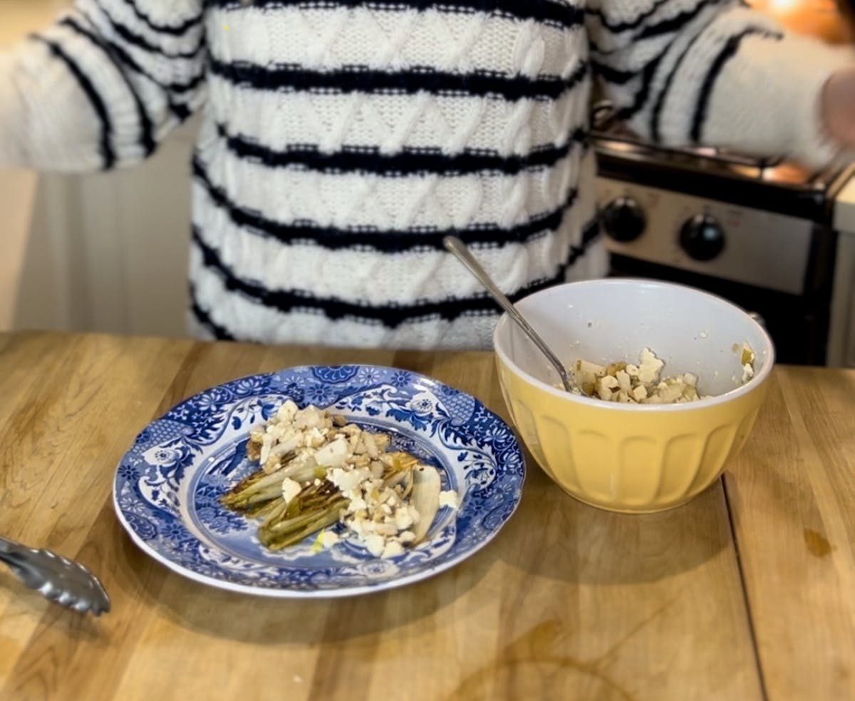 a grillled endive salad on a blue plate next to a yellow bowl with someone in a black and white striped sweater standing behind
