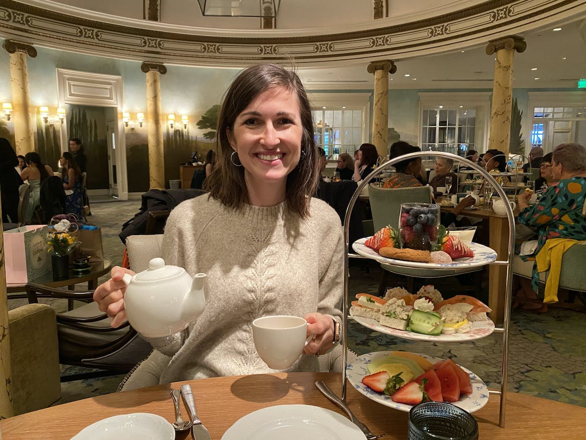 woman smiling with a 3 tiered tray of fruit and pastries and holding a pot of tea